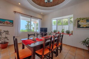 A dining area with a wooden table, six chairs, and a TV. There are windows with curtains, potted plants, and artwork on the walls.