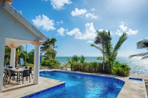 A pool area with a view of the ocean, surrounded by palm trees and a covered seating area with chairs and a table.