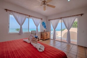 A bedroom with a large bed, two windows with curtains, a ceiling fan, and a view of the ocean.