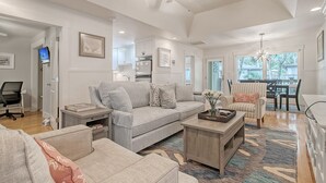 A spacious living room with a grey sofa, a coffee table, and a striped armchair. A kitchen is visible in the background, and a dining area with a table and chairs is also seen. - Completely Renovated, 5 min walk to beach (Kiawah Island)
