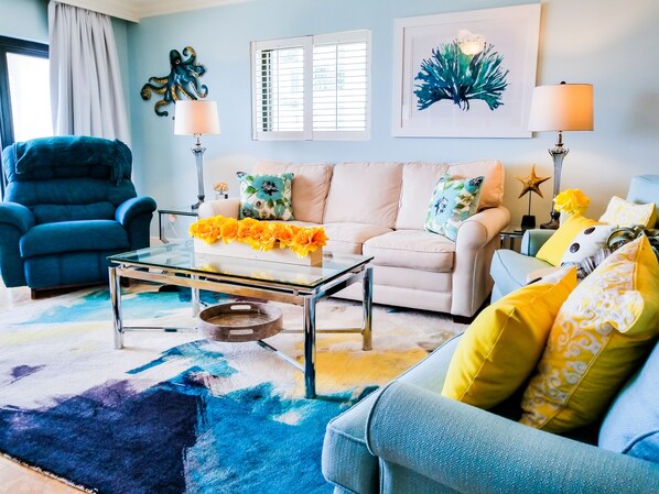 Un salon avec un fauteuil bleu, un canapé blanc orné de coussins à motifs floraux, une table basse en verre et un tapis coloré.