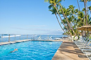 A swimming pool with a person swimming, surrounded by lounge chairs and palm trees, with a view of the ocean and a mountain in the background. - 139-2 Hale Aloe Great spot to spend a vacation in the sun!! (Lahaina)