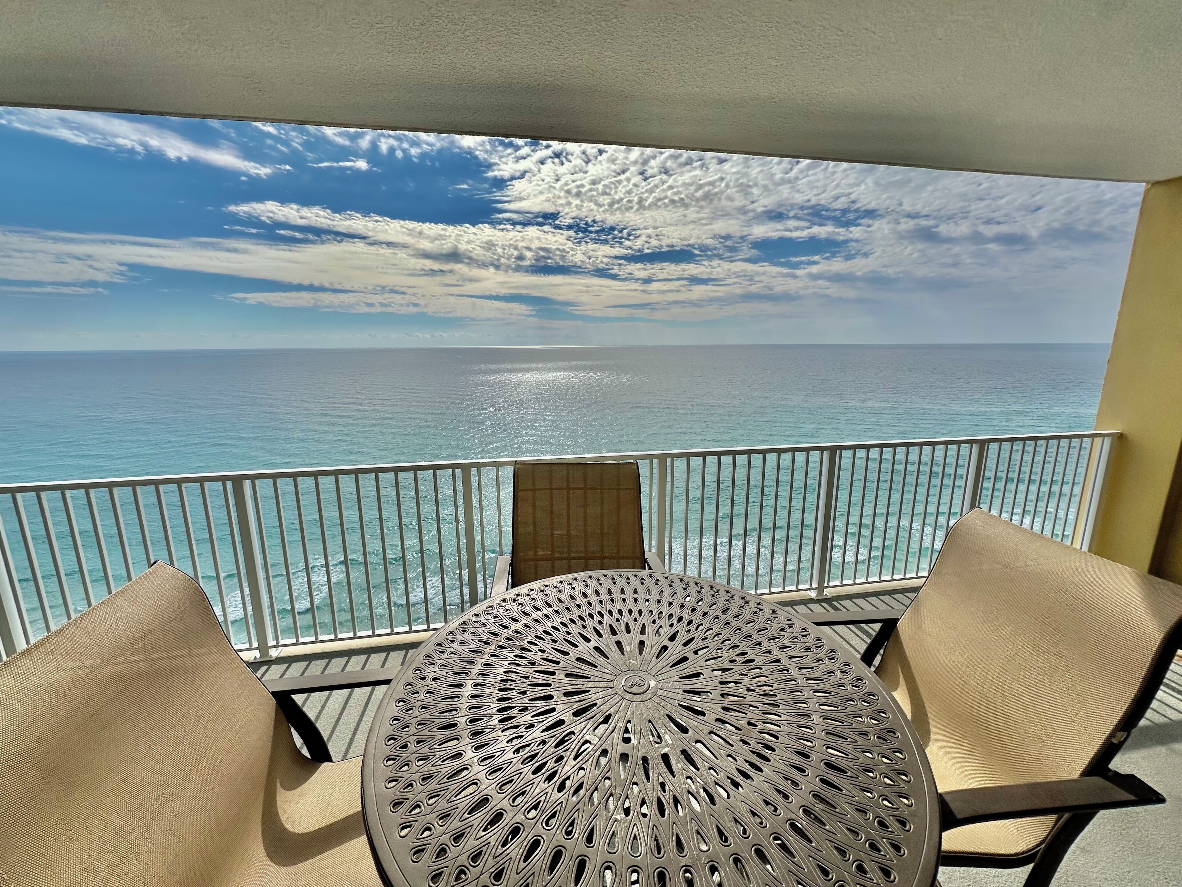 A balcony with a round table and two chairs, overlooking a turquoise sea and a clear sky.