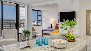 Una habitación de hotel moderna con un ventanal grande, una cama, un televisor, una zona de comedor con encimera de mármol y un balcón con vistas a la ciudad.