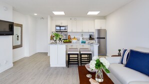 Una cocina moderna con armarios blancos, microondas integrado y refrigerador de acero inoxidable. Hay una pequeña isla con taburetes de bar y un florero con flores.