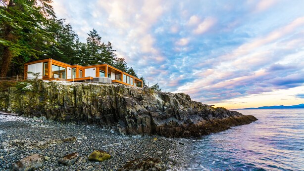 A modern coastal house with large windows, situated on a rocky shoreline with a view of the sea and mountains in the background. - UNIQUE RESIDENTIAL OCEANFRONT PROPERTY ON VANCOUVER ISLAND  (Nanoose Bay)