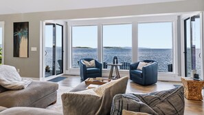 A modern living room with a large window overlooking a body of water, two blue armchairs, a grey sofa, and a small table with a tray.