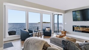 A modern living room with a large window overlooking a body of water, a fireplace, and a flat-screen TV mounted on the wall. - UNIQUE RESIDENTIAL OCEANFRONT PROPERTY ON VANCOUVER ISLAND (Nanoose Bay)