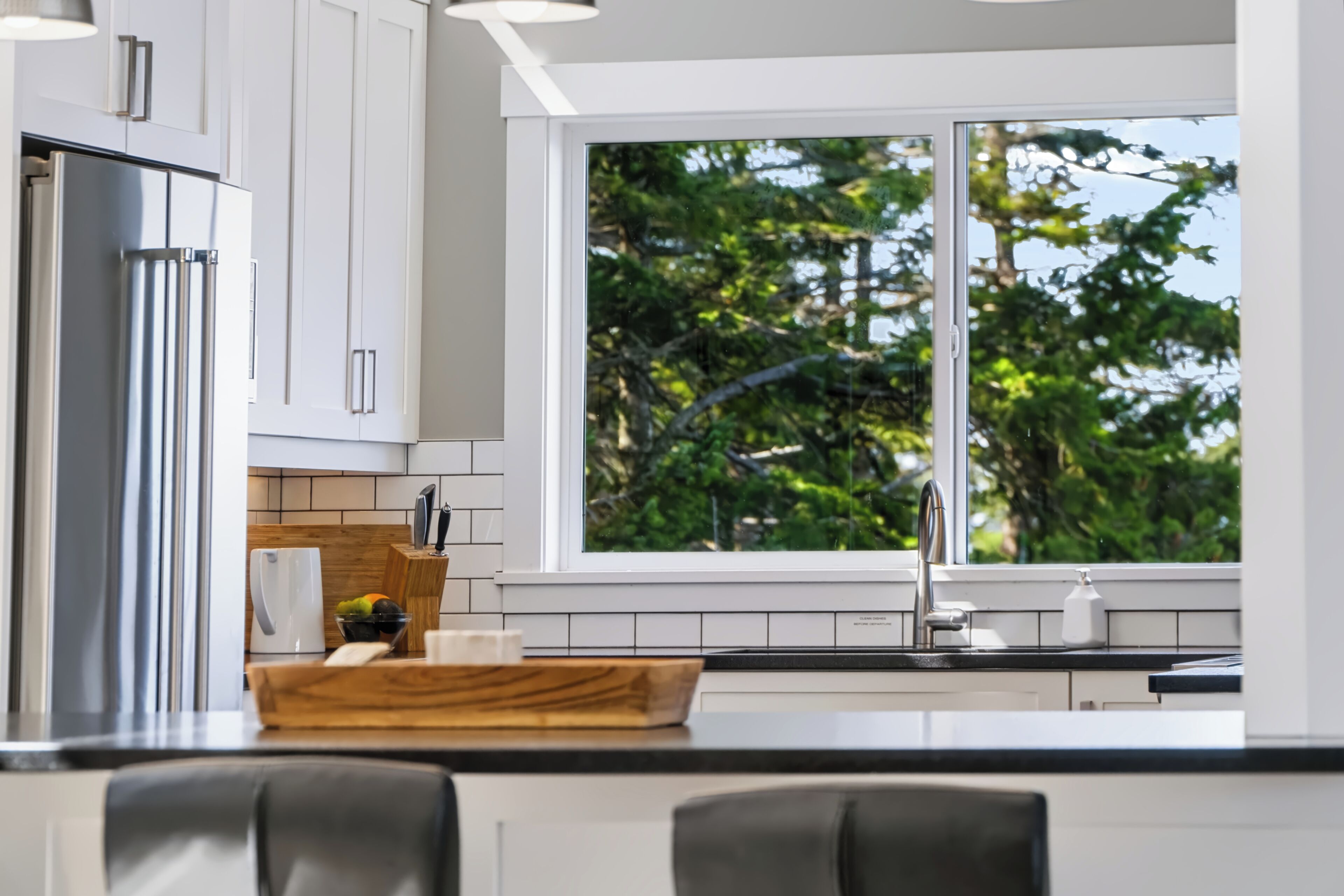 A modern kitchen with white cabinets, a stainless steel refrigerator, a wooden cutting board, and a window with a view of trees.