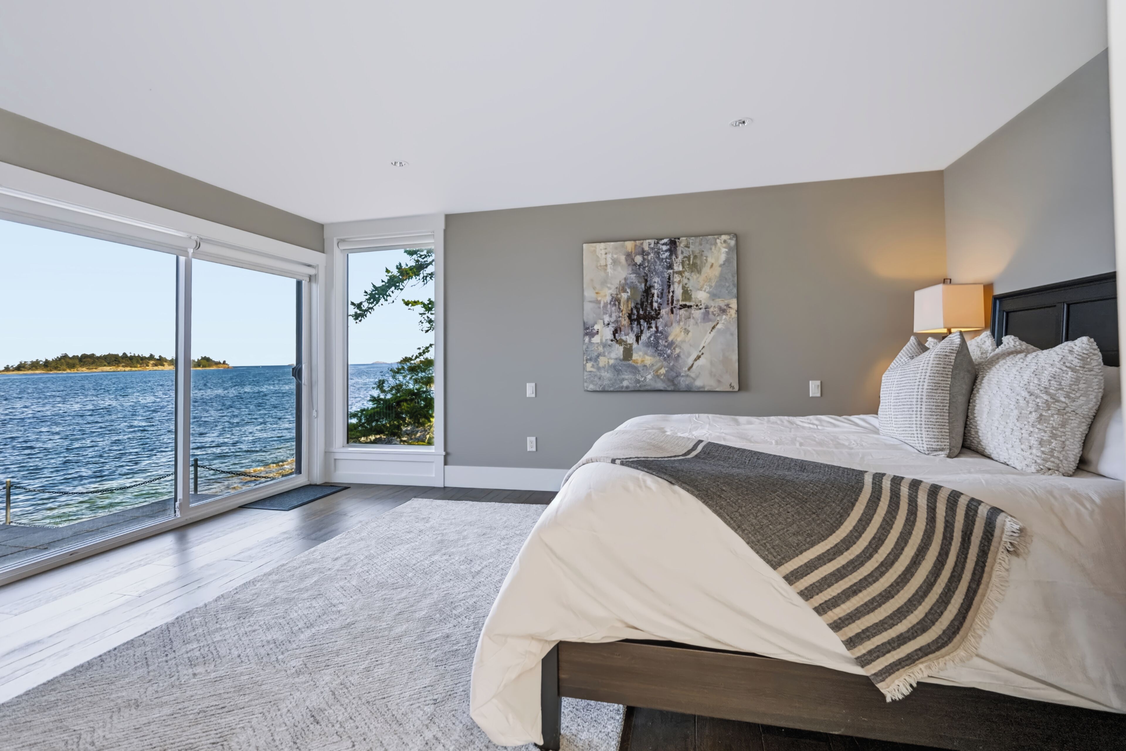 A bedroom with a large bed, a painting, and a view of the sea.