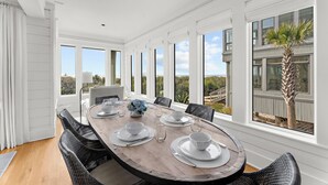 A dining area with a large wooden table set for six, surrounded by wicker chairs, with a view of the outdoors through large windows. - Incredible, completely renovated oceanfront villa (Kiawah Island)