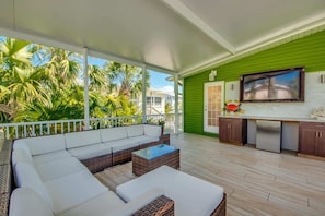 A covered outdoor area with a white sofa, wicker coffee table, and a kitchen with a TV. - 275 Ohio Charming Pool Home near the Beach (Fort Myers Beach)