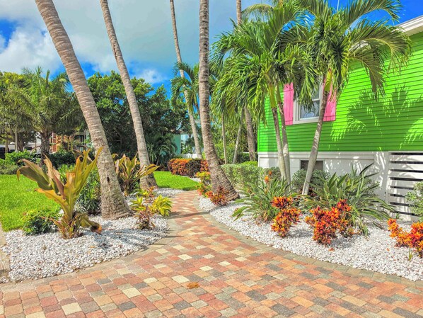 A pathway leading to a green house with a pink window, surrounded by palm trees and tropical plants.