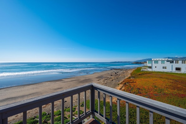 Vista costera desde un balcón con vistas a una playa y al océano, con una casa moderna al fondo.