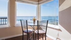 Een hoekappartement met twee grote ramen met uitzicht op de oceaan, een ronde tafel met stoelen en een vaas met bloemen.