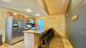 A compact kitchen with wooden cabinets, a stainless steel refrigerator, a microwave, and a sink. There is a wooden island with stools and a window providing natural light. - Beautiful mountain cabin (Sevierville)