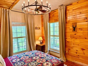 Une chambre avec un mur en bois, un lit recouvert d’une courtepointe aux couleurs vives, un lustre et une fenêtre avec des rideaux.