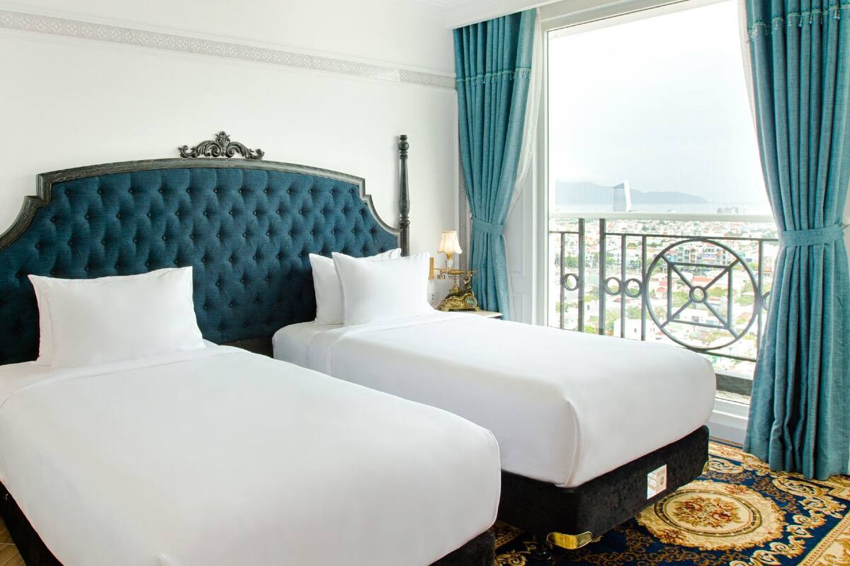 A hotel room with two beds, a large window with a view of the city, a nightstand with a lamp, and a decorative headboard.