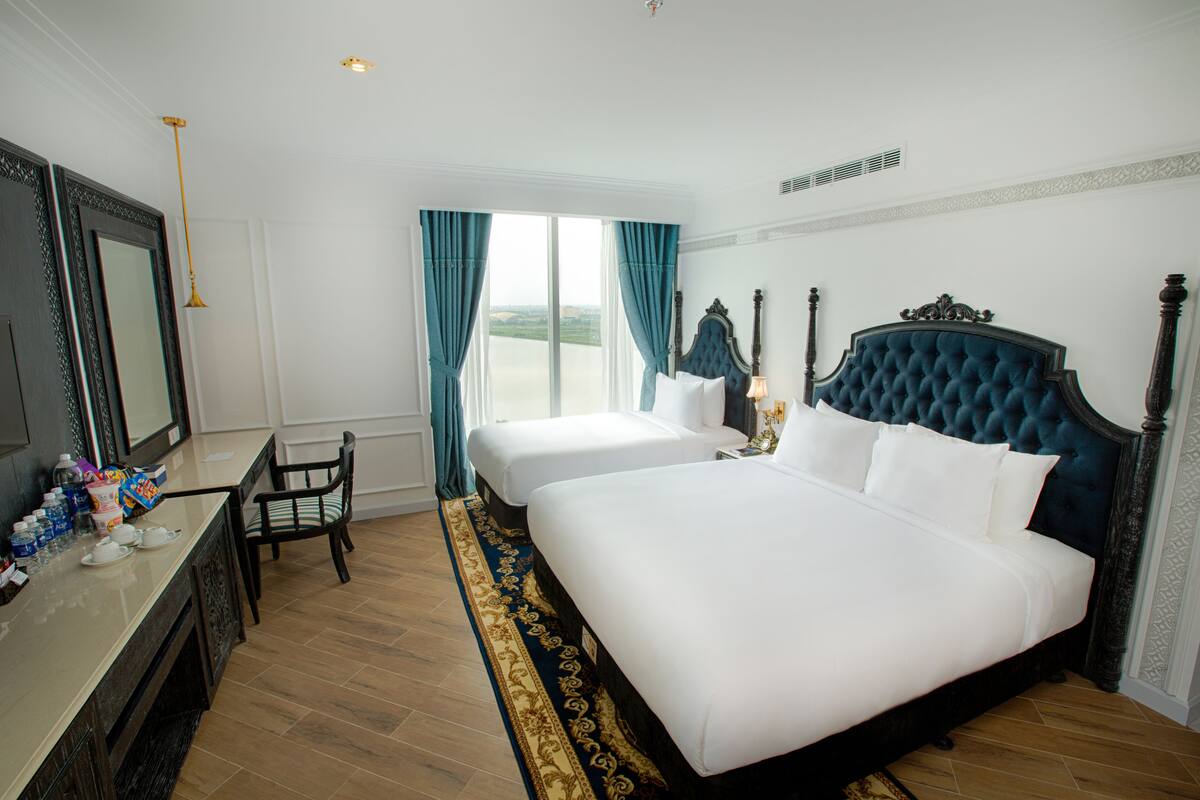 A hotel room with two beds, a desk with a chair, a large window with curtains, and a view of the outdoors.