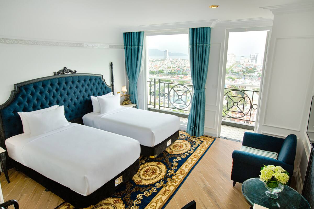 A hotel room with two beds, a balcony with a city view, and a small table with a vase of flowers.