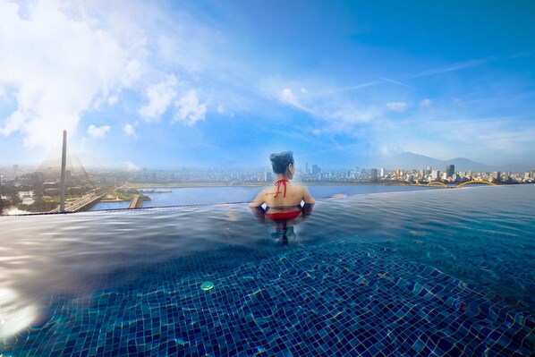 A person sitting on an infinity pool with a cityscape and a bridge in the background. - Fivitel Da Nang (Da Nang)