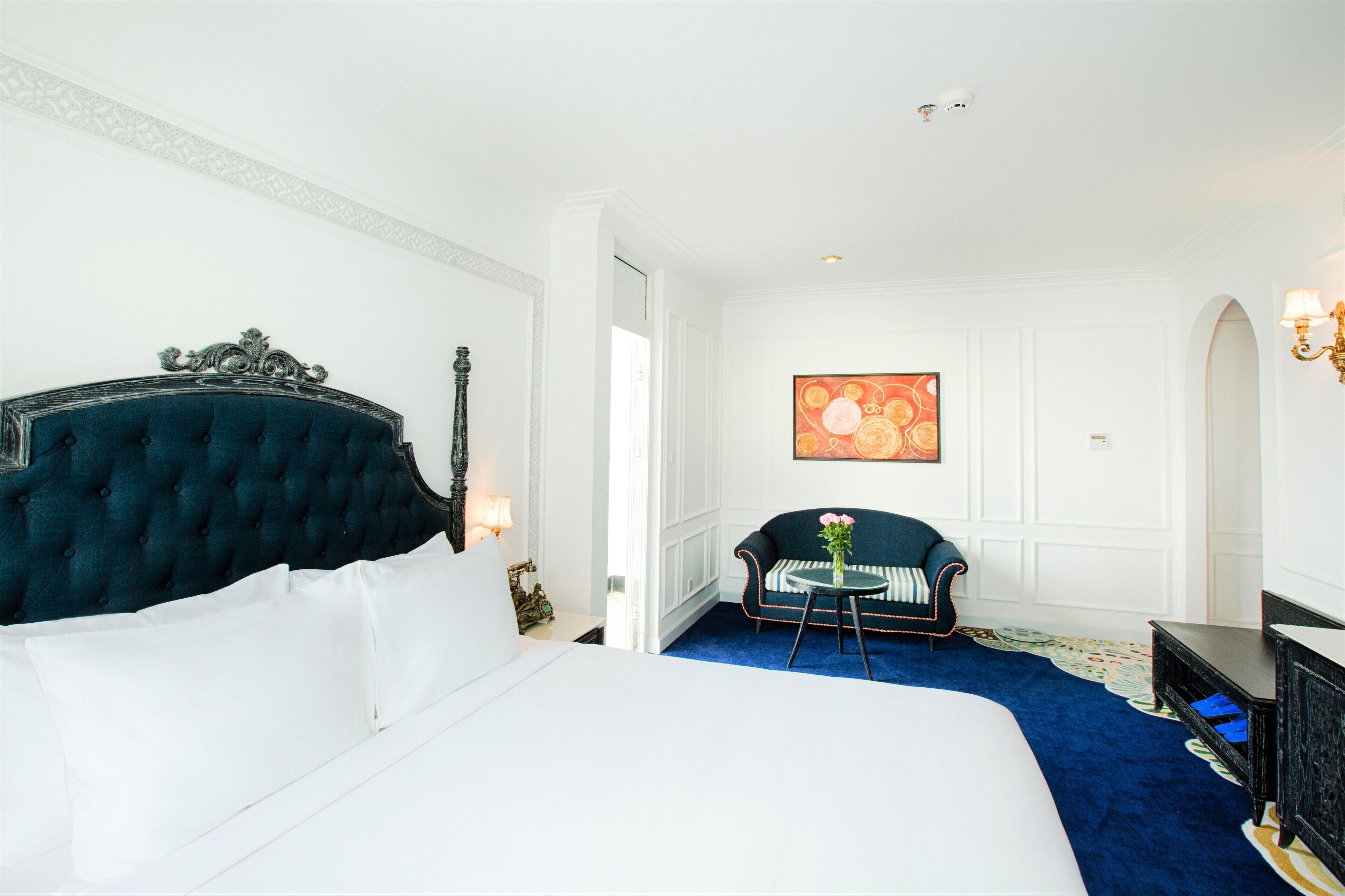 A hotel room with a large bed, a dark upholstered headboard, a sofa, a small round table with a vase, and a painting on the wall.