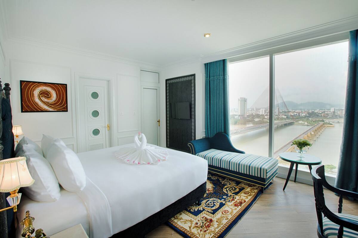 A hotel room with a large bed, a seating area, a view of a bridge and cityscape, and a large window with curtains.