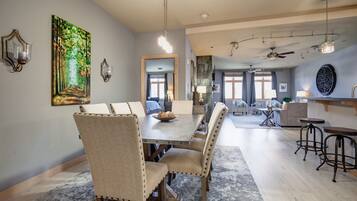 A spacious dining area with a long table and chairs, a painting on the wall, and a view into the kitchen.