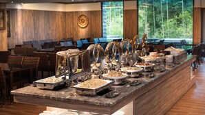 En buffet med forskellige retter, et siddeområde med stole og borde samt et stort vindue med udsigt over grøn vegetation.