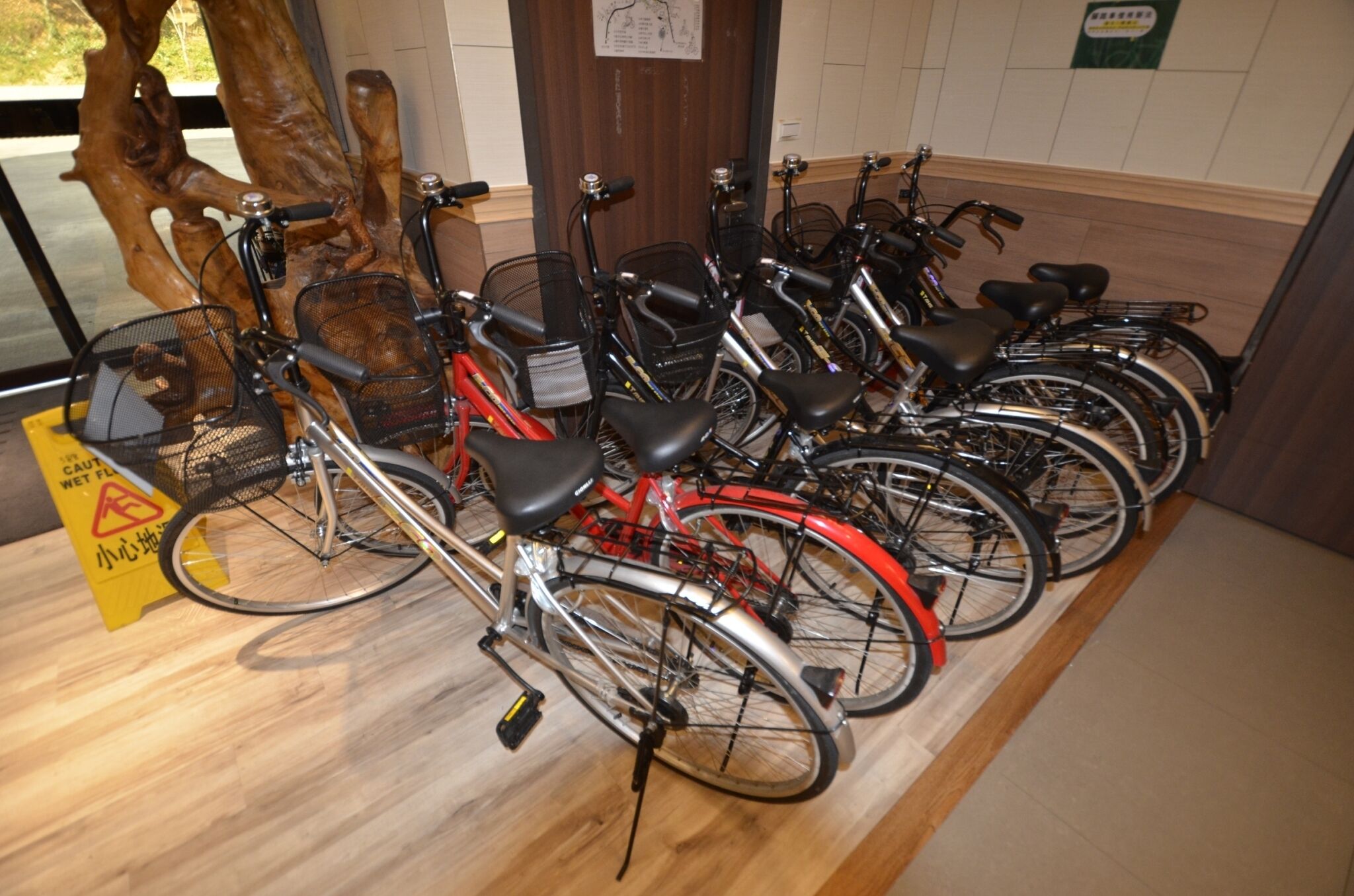 A row of bicycles parked indoors, with a wooden floor and a caution sign visible.