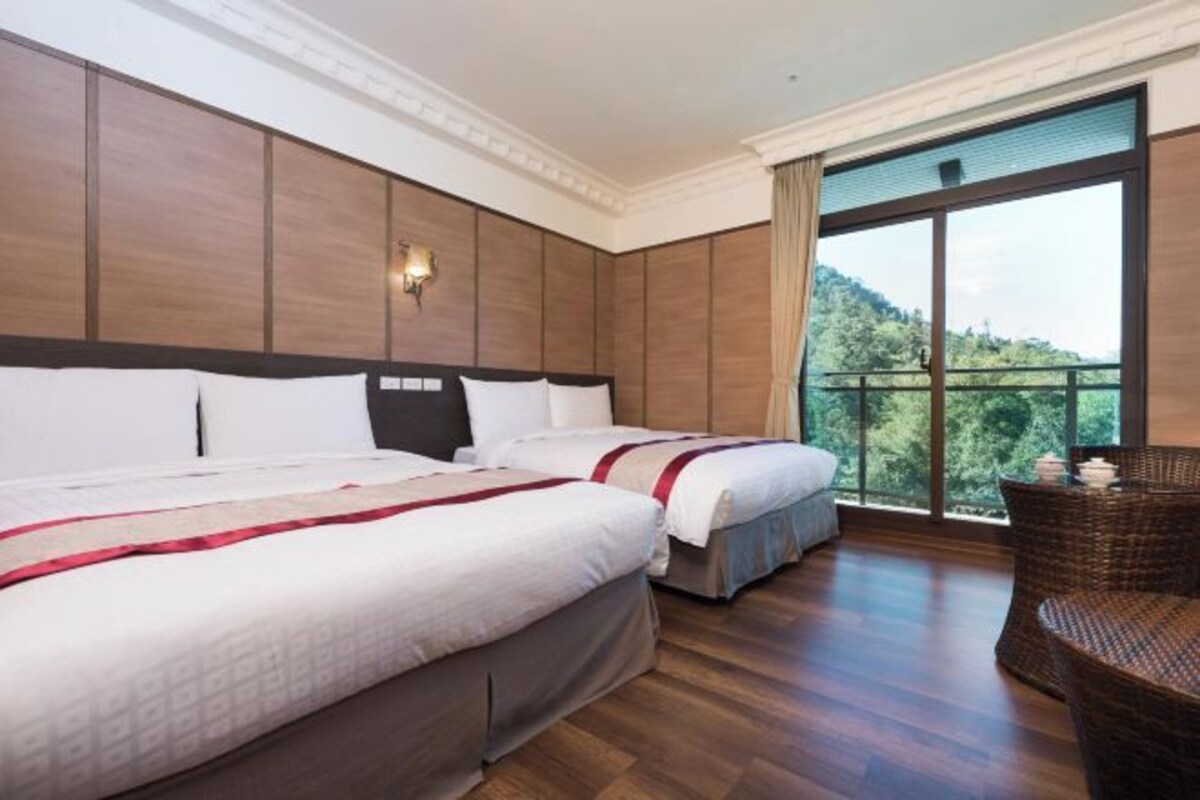 A hotel room with two beds, a large window with a view of greenery, and a wicker chair.