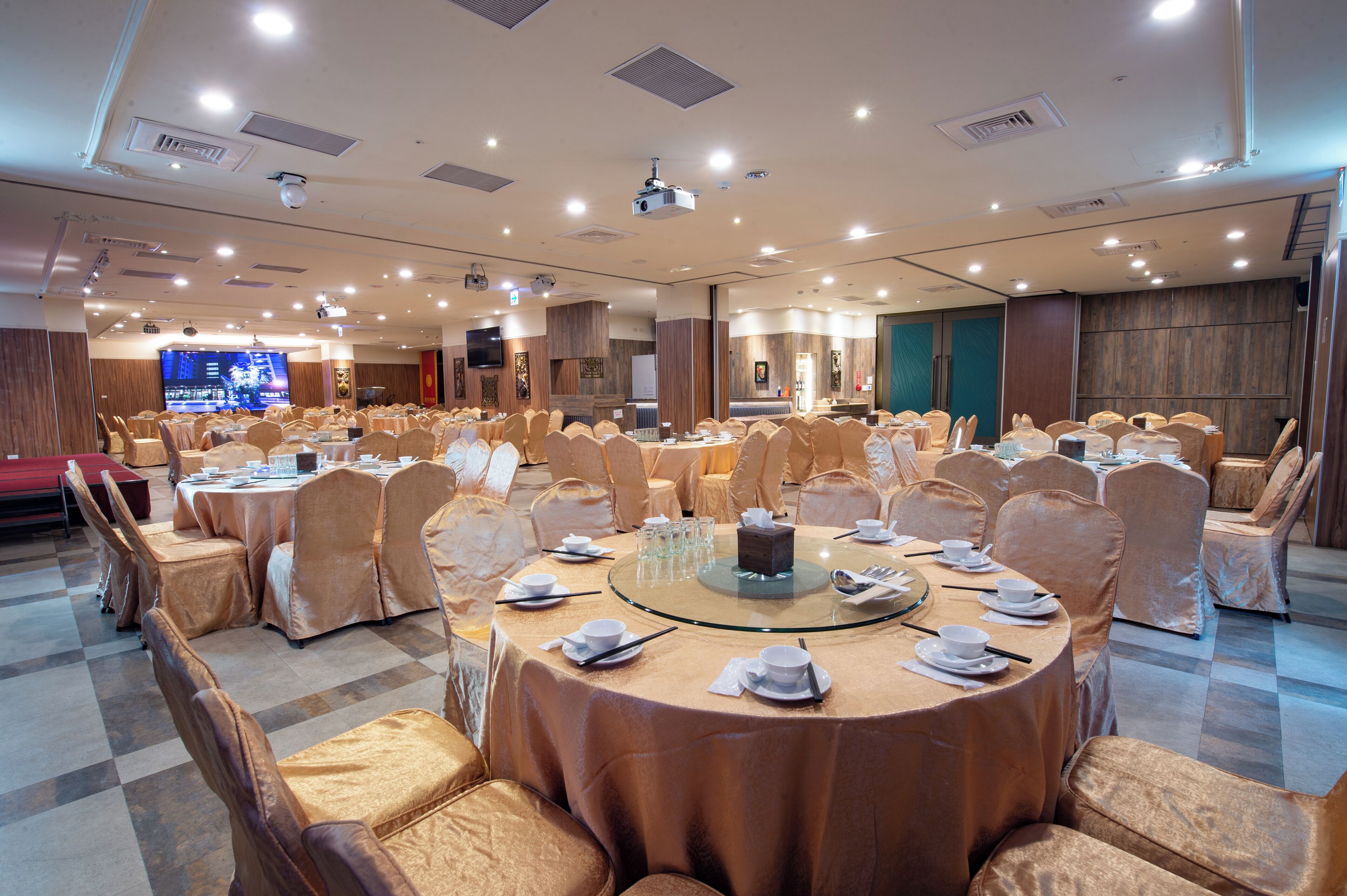 Un salón de banquetes con mesas redondas, sillas cubiertas y un escenario con una pantalla grande.
