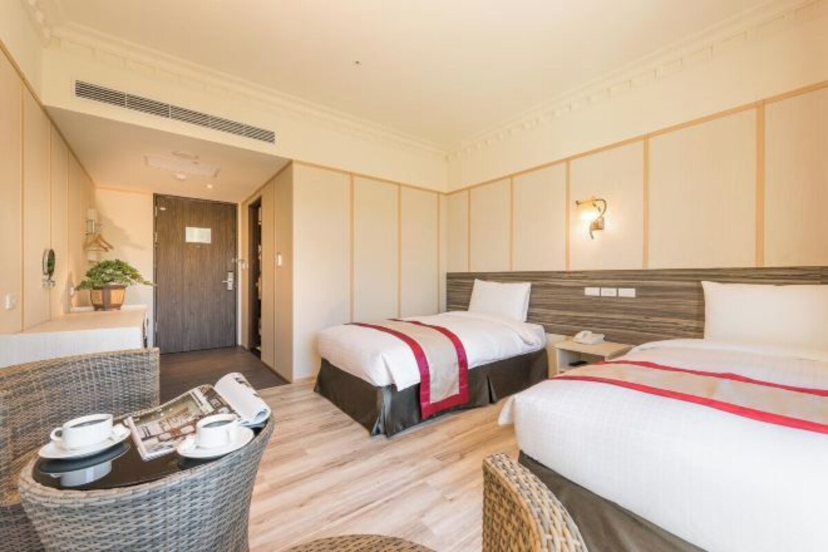 A hotel room with two beds, a small dining table, a chair, and a bedside table with a lamp.