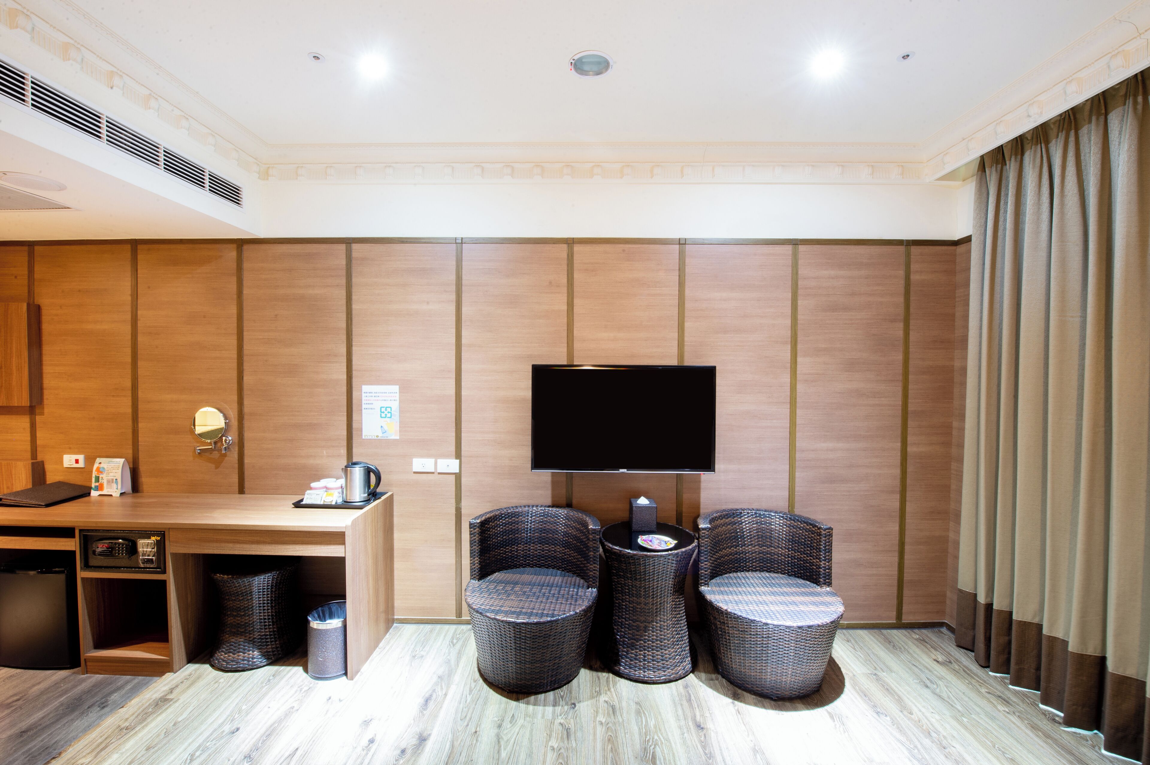 A hotel room with a wooden panel wall, a flat-screen TV, a desk with a coffee maker, two wicker chairs, and a trash can.