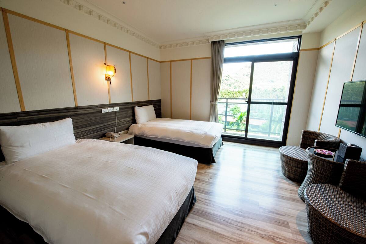 Habitación de hotel con dos camas, un ventanal grande, televisión y muebles de mimbre.