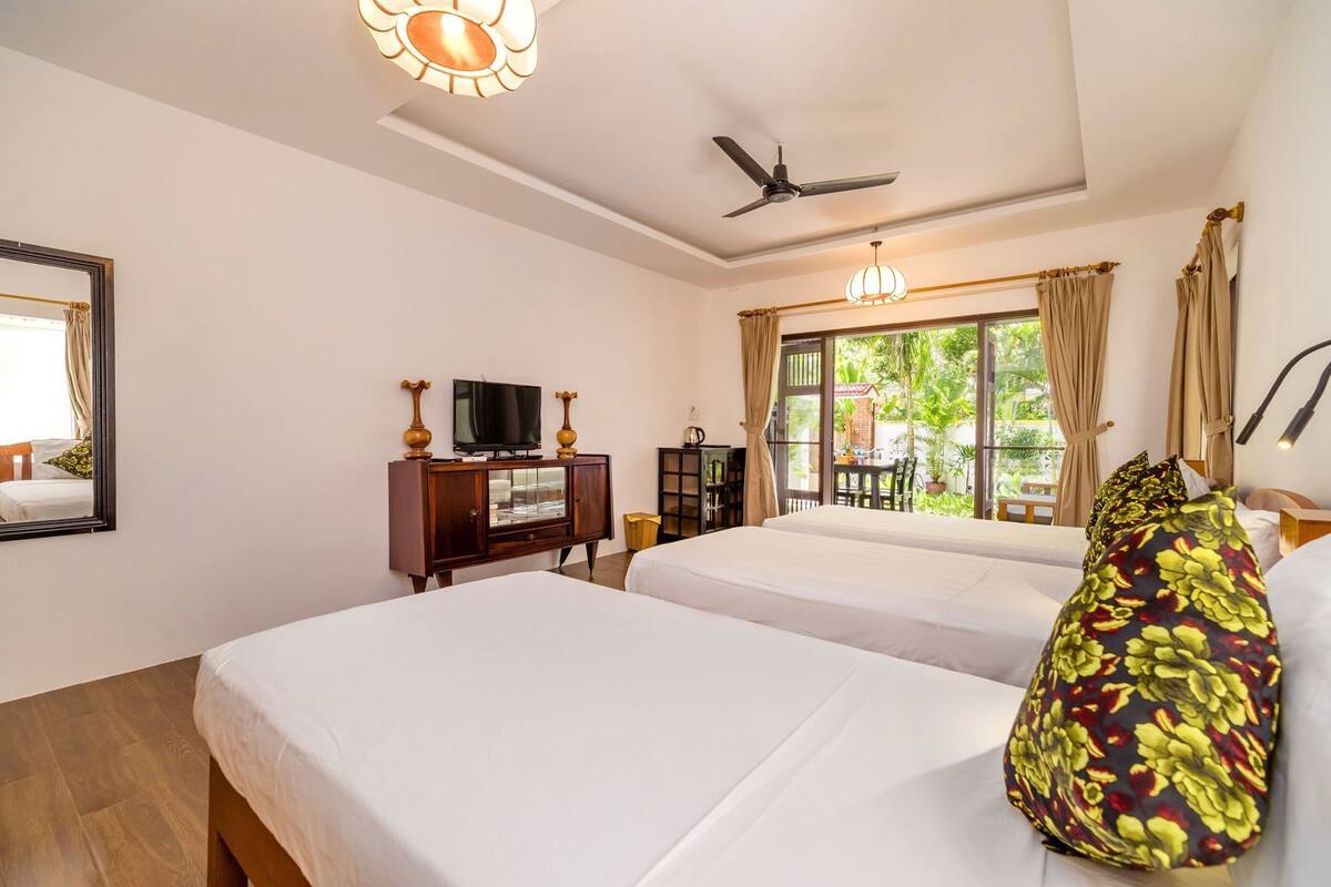 A hotel room with two beds, a TV, a ceiling fan, and a view of greenery outside.