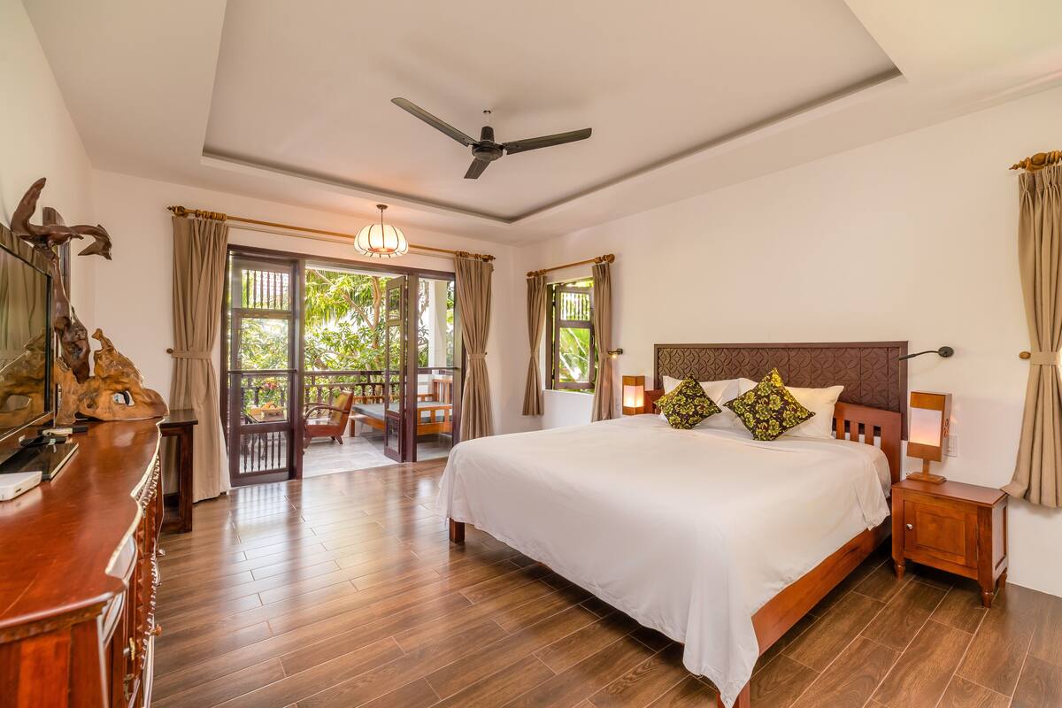A spacious bedroom with a large bed, wooden floor, ceiling fan, and a view of the outdoors.