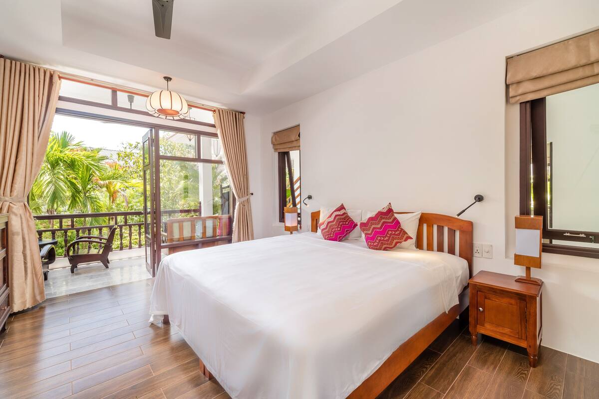A bedroom with a large bed, wooden nightstand, and a view of greenery outside.