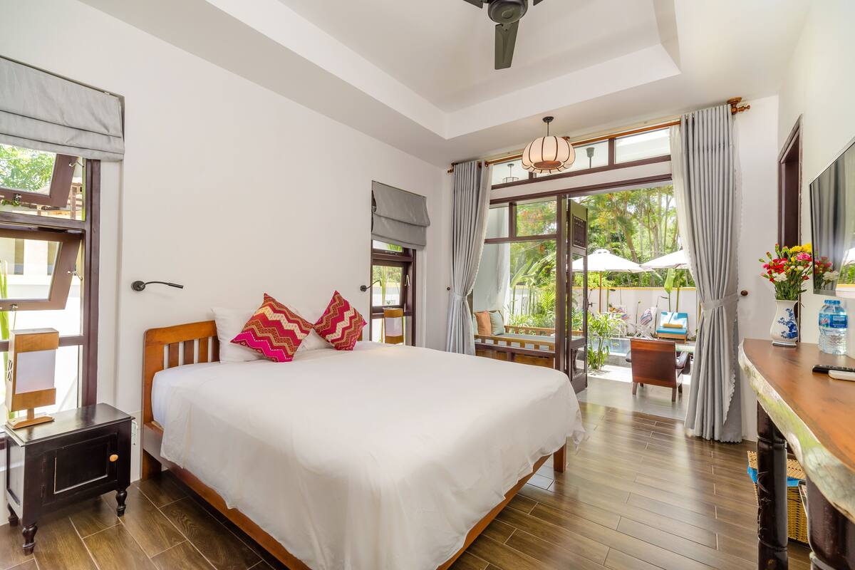 A hotel room with a large bed, wooden flooring, a ceiling fan, and a view of the outdoors through large windows.
