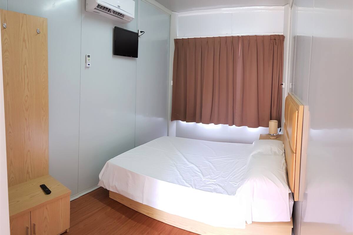 Un lit simple avec une tête de lit en bois, une table de chevet avec une lampe, une armoire et un climatiseur fixé au mur.
