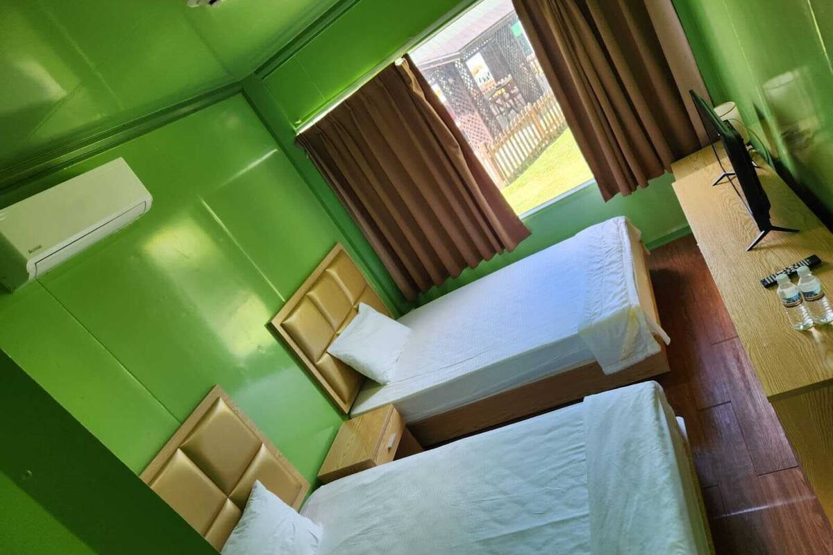 Une chambre avec deux lits, un bureau en bois, une chaise, une fenêtre avec des rideaux et une vue sur l’extérieur.