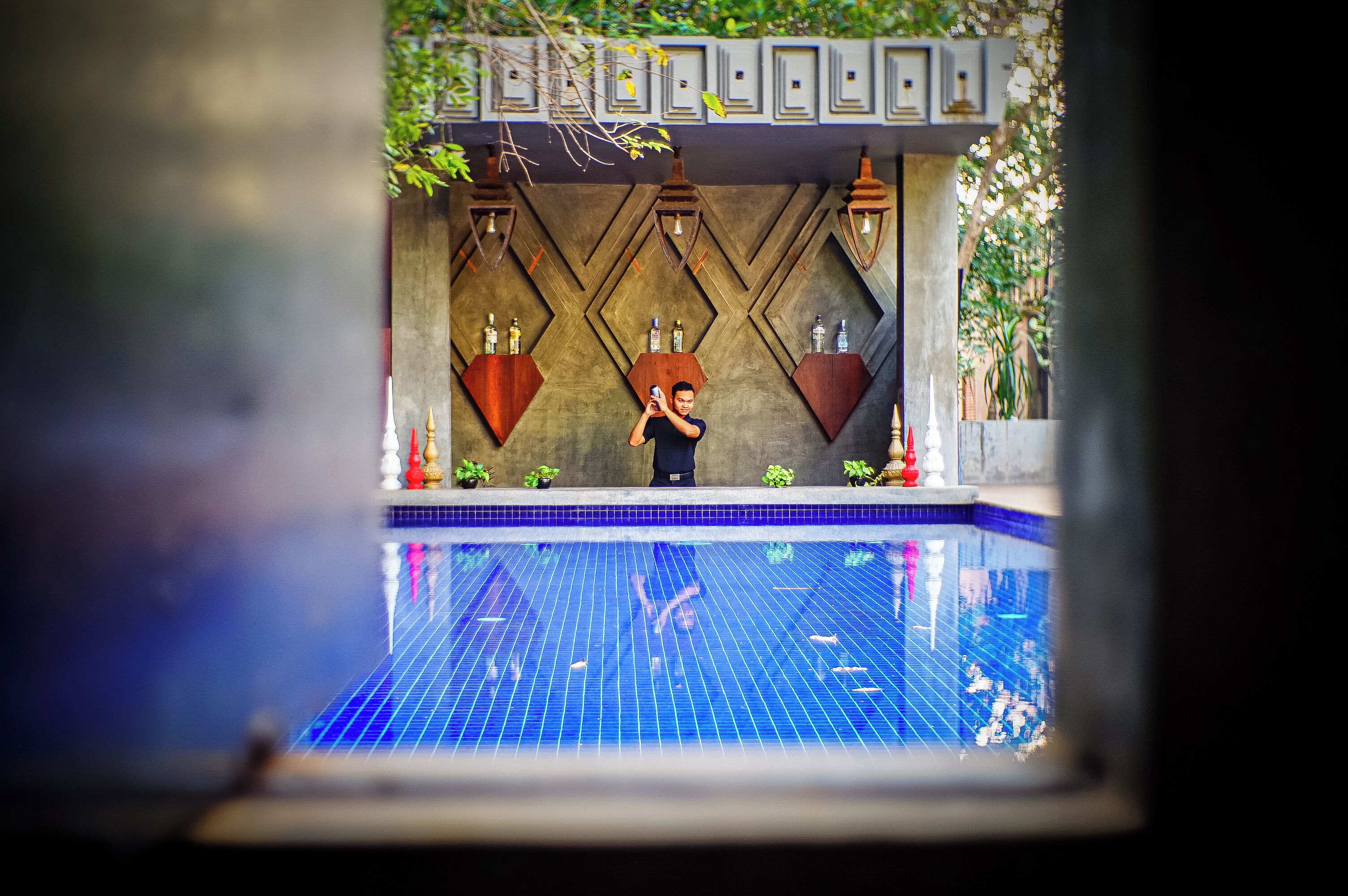 Un espace piscine avec un sol carrelé, une personne prenant une photo et des éléments décoratifs tels que des plantes et des lampes fixées au mur.