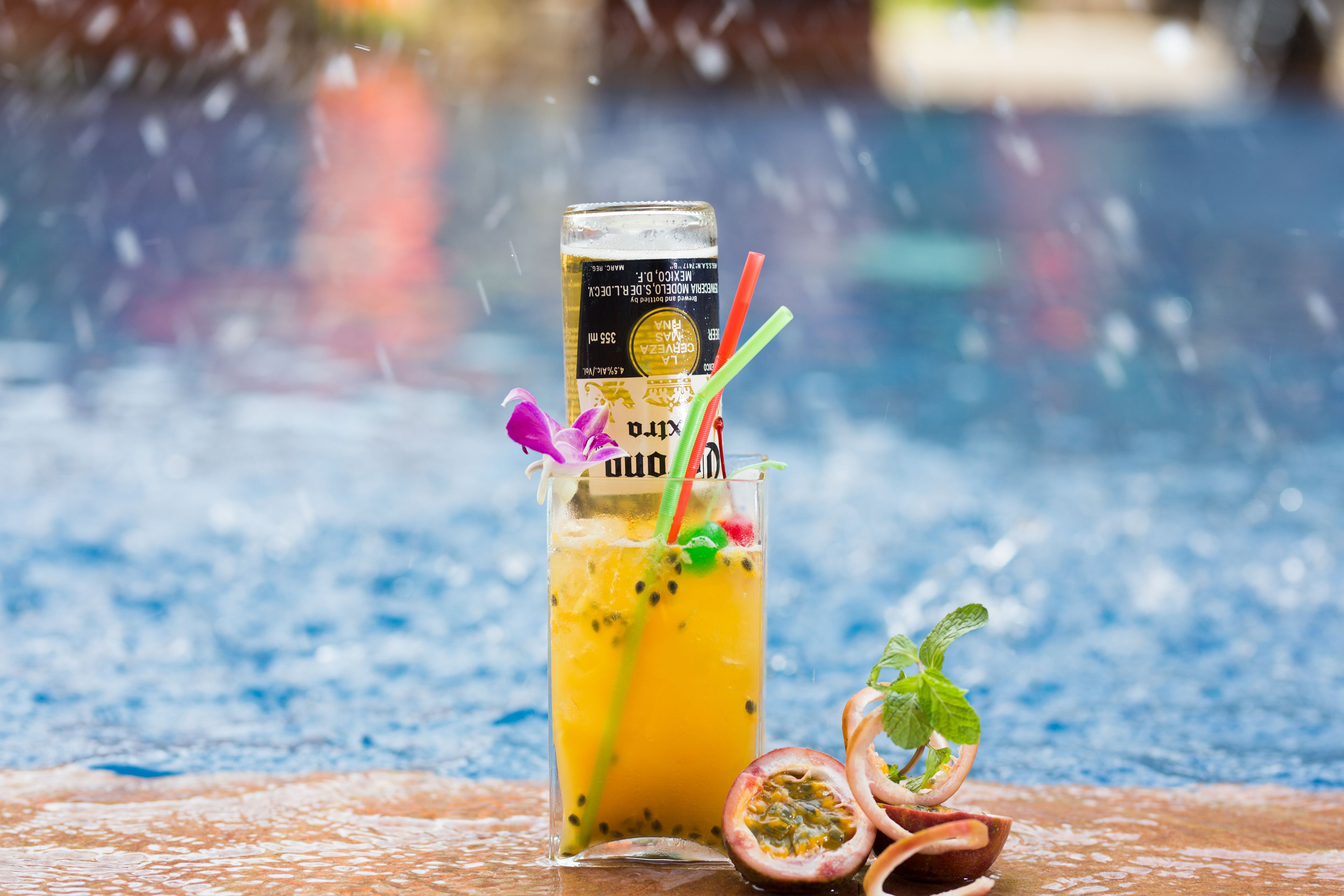 A glass of tropical drink with a can of beer, a straw, and a slice of passion fruit on a textured surface.