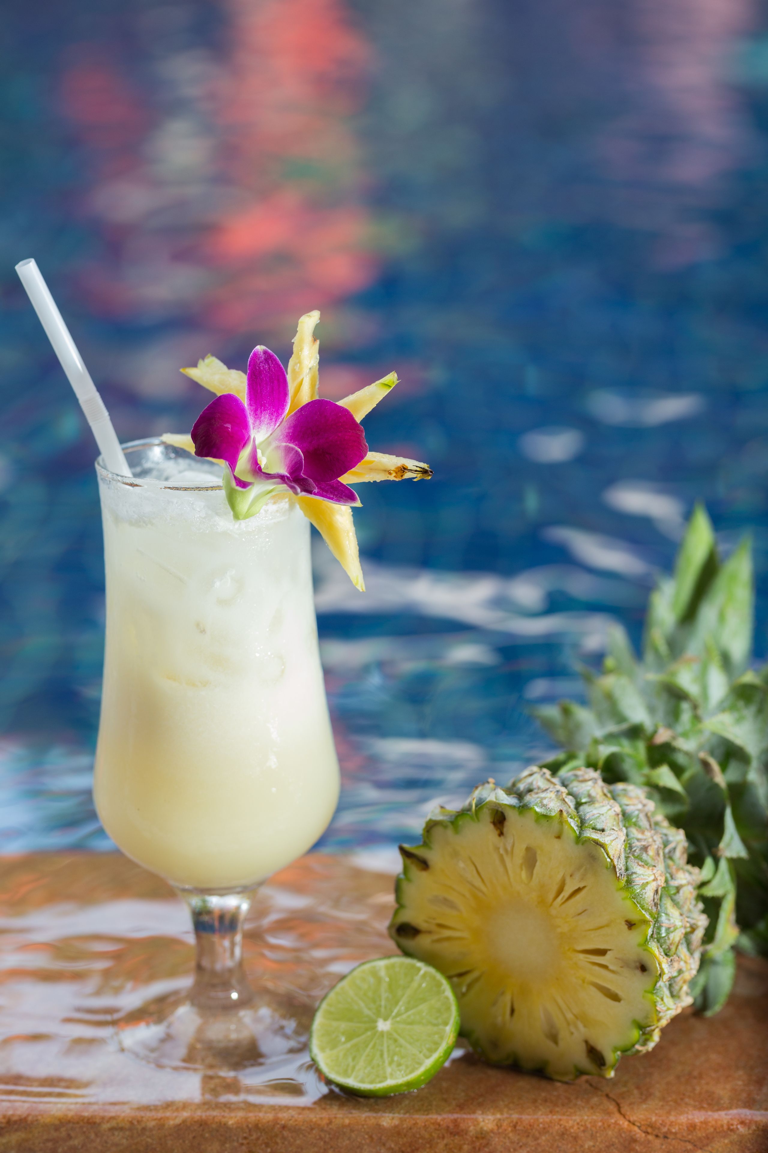 A glass of a light-colored beverage with a straw, garnished with a purple flower, next to a pineapple and a lime half on a wooden surface.