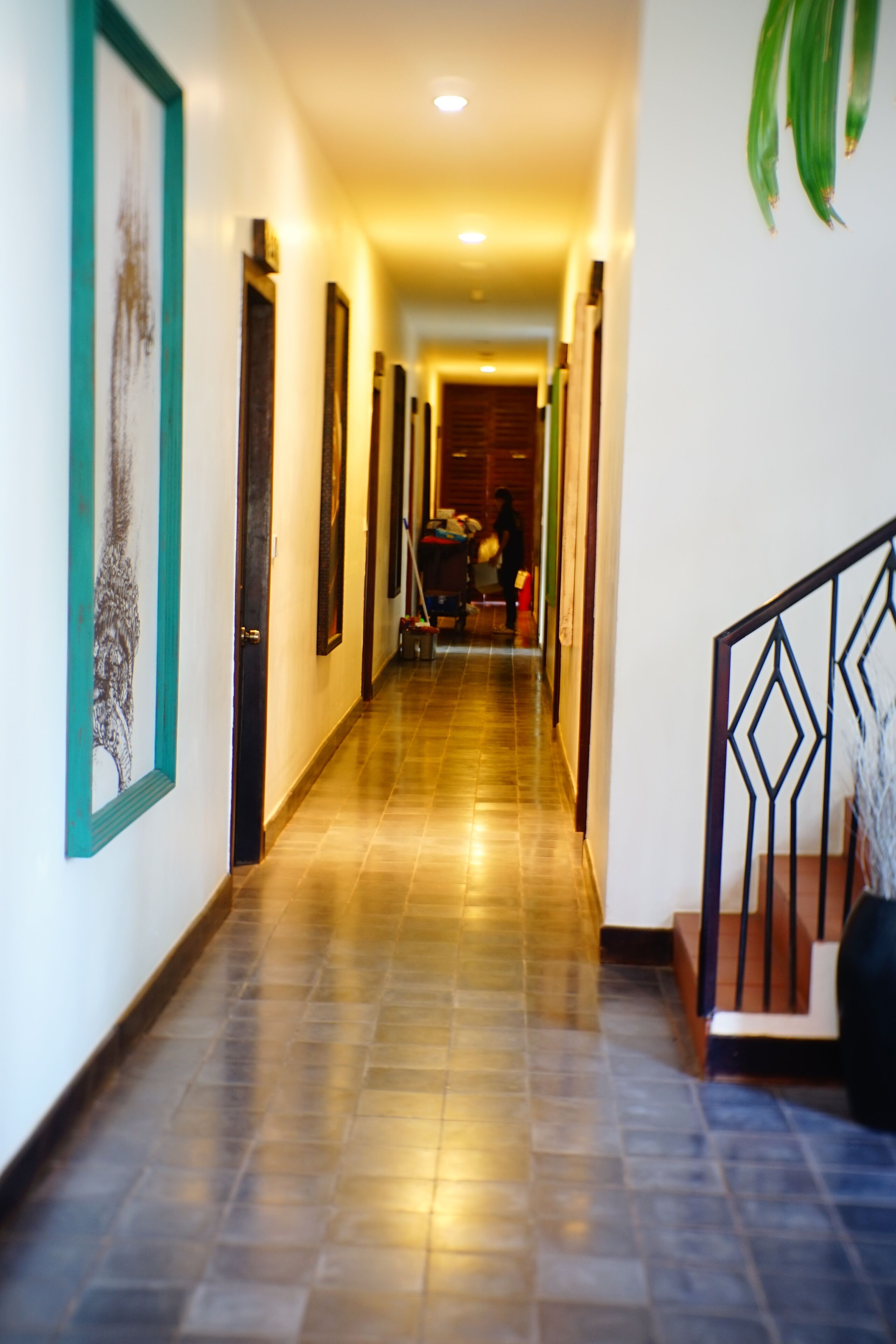 Un pasillo con suelo de baldosas, obras de arte enmarcadas en las paredes y una escalera con barandilla geométrica.