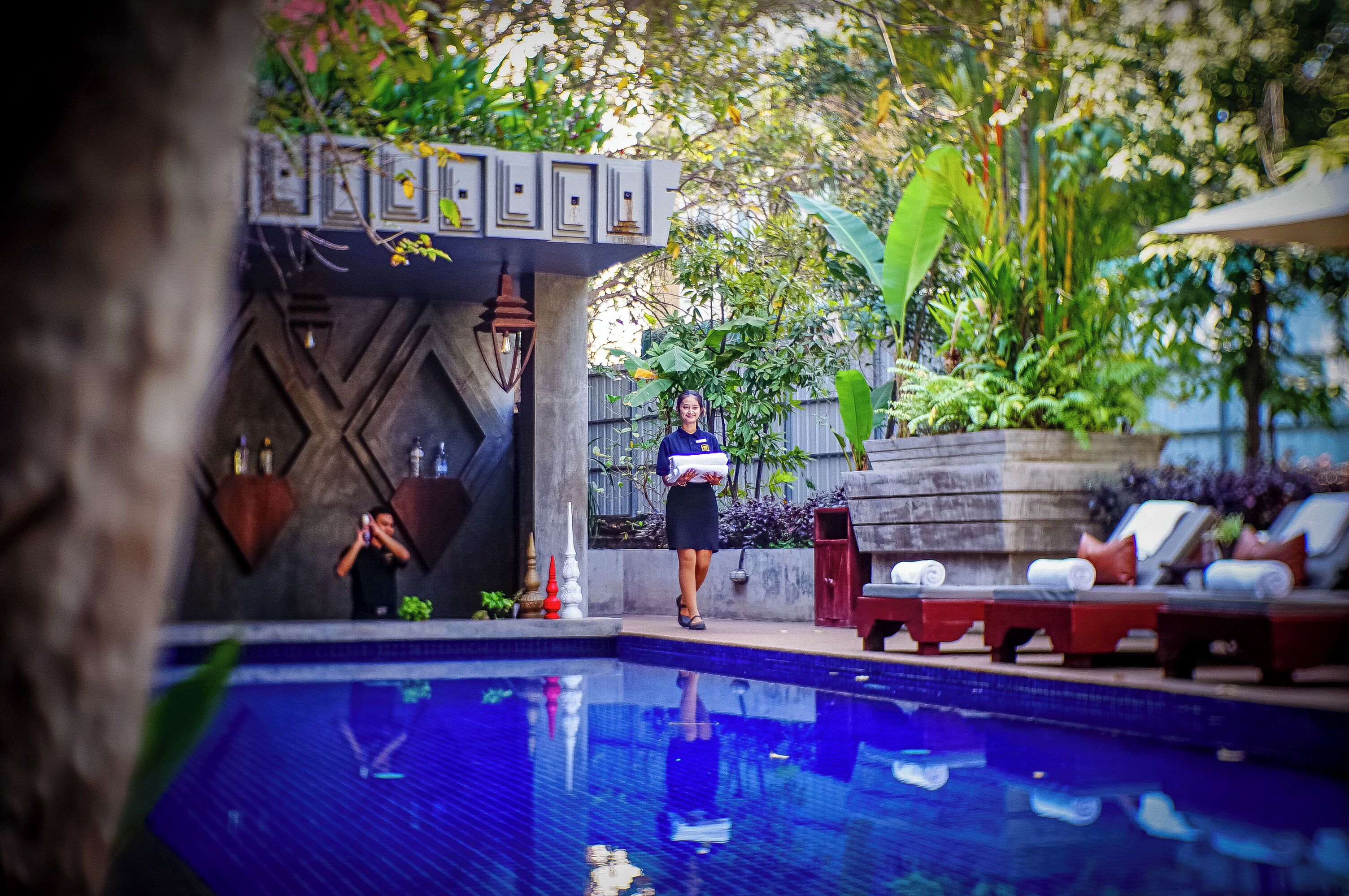 Un área junto a la piscina con sillones de descanso, una persona sosteniendo una toalla y una pared decorativa con diseños geométricos.