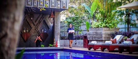 Un área junto a la piscina con sillones de descanso, una persona sosteniendo una toalla y una pared decorativa con diseños geométricos.