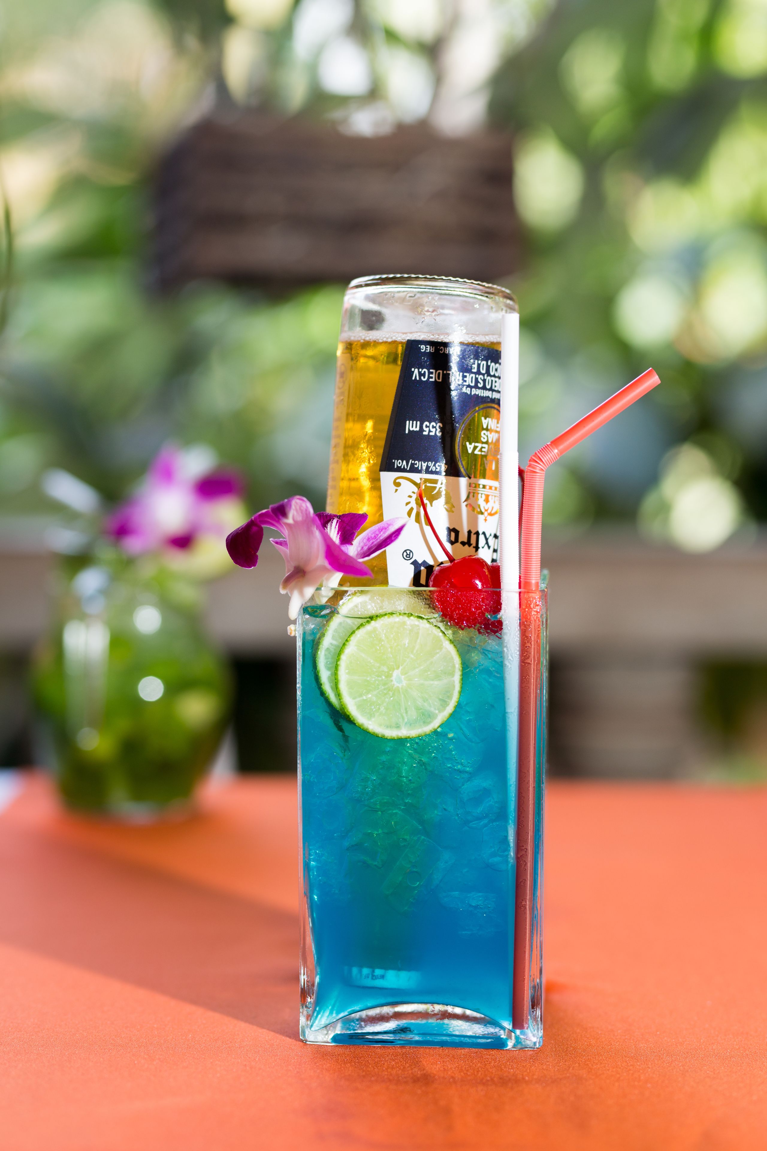 Un vaso alto con una bebida azul, una rodaja de lima, una cereza y una pajita, sobre una mesa con un fondo verde desenfocado.