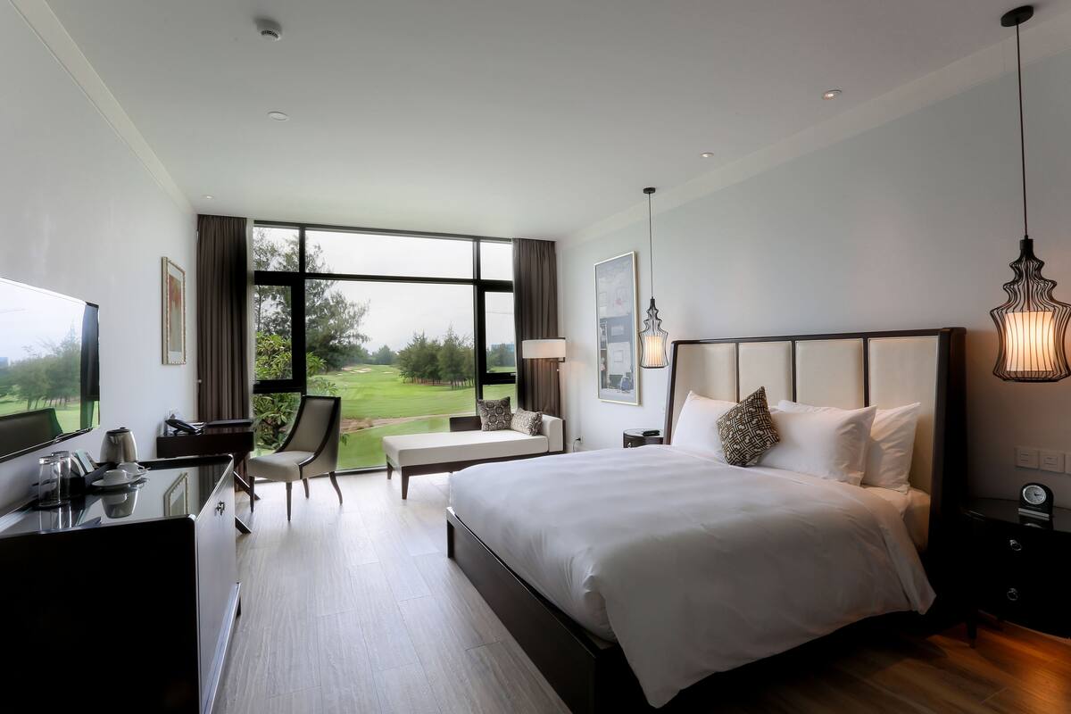 Moderni hotellihuone, jossa on suuri sänky, työpöytä, tuoli ja näkymä golfkentälle.