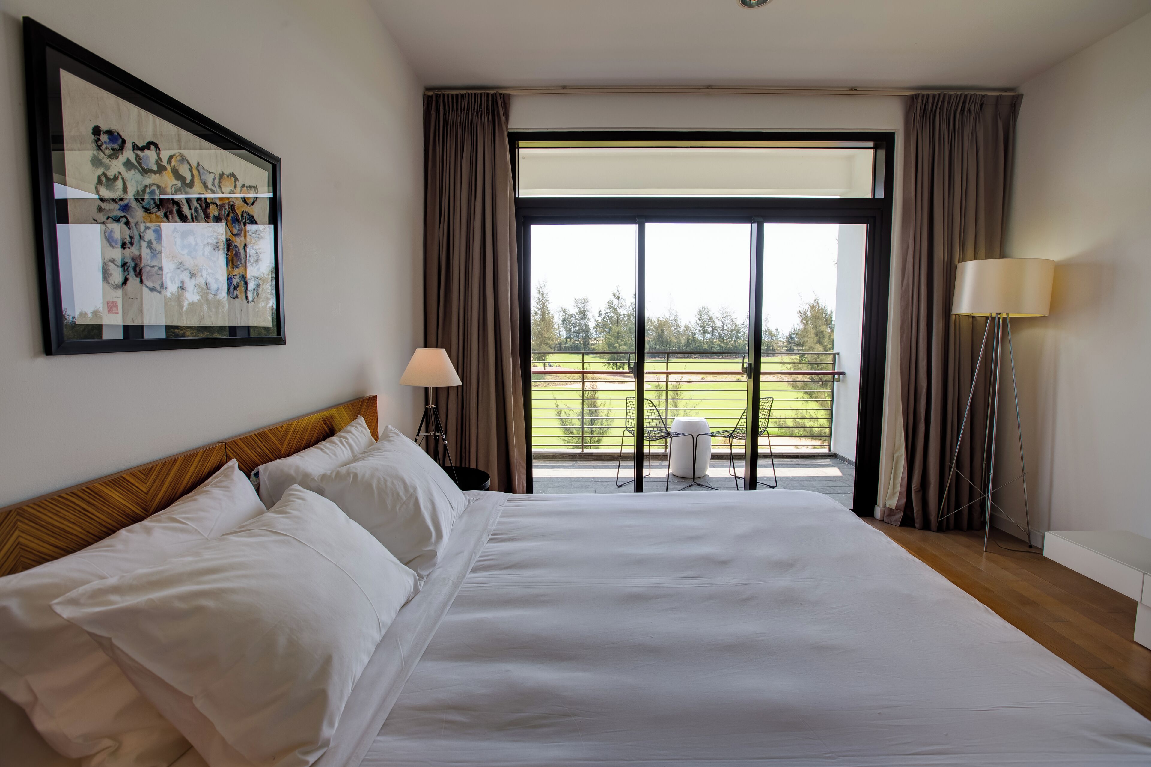 A hotel room with a large bed, a framed artwork on the wall, a balcony with a view of the outdoors, and a floor lamp.